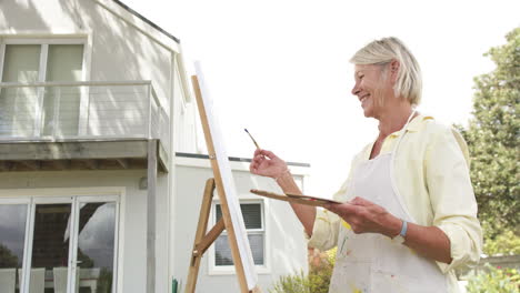 Happy-caucasian-senior-woman-painting-picture-and-smiling-in-sunny-garden,-slow-motion,-copy-space