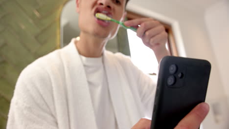 Biracial-man-brushing-teeth-and-using-smartphone-in-bathroom-in-the-morning,-slow-motion