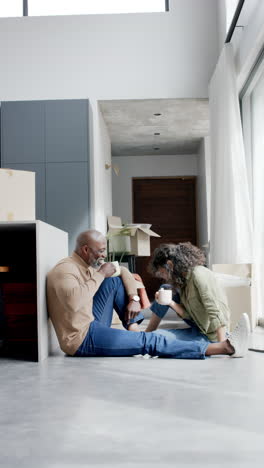 Vertical-video-of-happy-diverse-couple-sitting-on-floor-drinking-coffee,-copy-space,-slow-motion