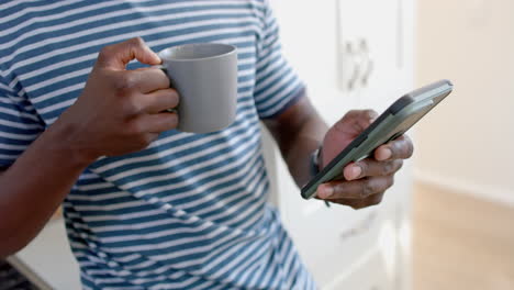 Glücklicher-Afroamerikanischer-Mann,-Der-Kaffee-Trinkt-Und-Sein-Smartphone-In-Einer-Sonnigen-Küche-Benutzt,-Zeitlupe