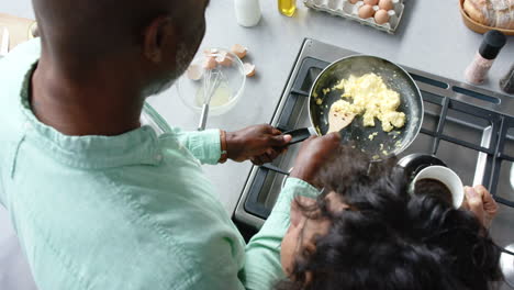 Happy-biracial-couple-cooking-and-preparing-scrambled-eggs-on-pan-in-kitchen,-slow-motion