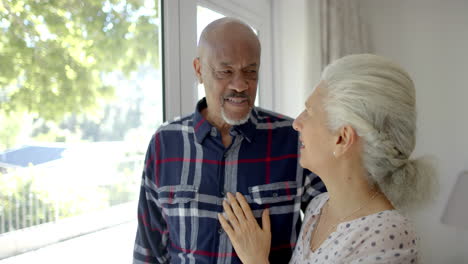 Happy-senior-biracial-couple-looking-through-window-and-talking-in-bedroom,-slow-motion