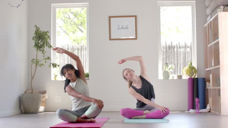 Happy-diverse-fitness-women-exercising-and-stretching-on-mat-in-white-room,-slow-motion