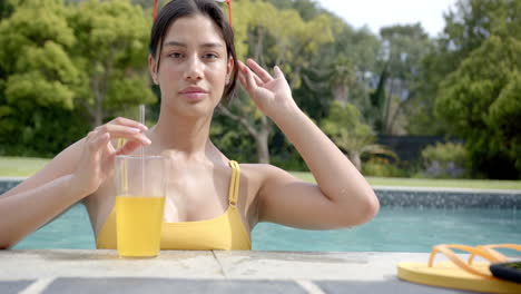 Portrait-of-happy-biracial-teenage-girl-standing-in-sunny-swimming-pool-with-juice,-slow-motion