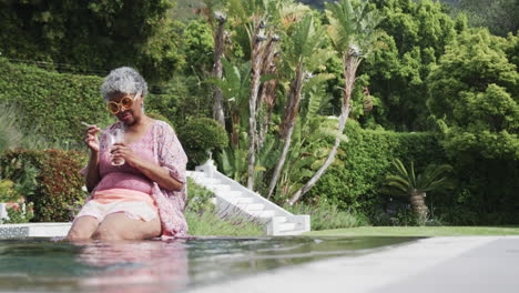 Happy-senior-african-american-woman-sitting-with-legs-in-pool-holding-drink,-copy-space,-slow-motion