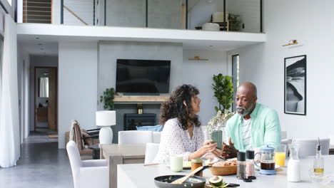 Happy-biracial-couple-having-breakfast-using-smartphone-and-talking-in-kitchen,-slow-motion