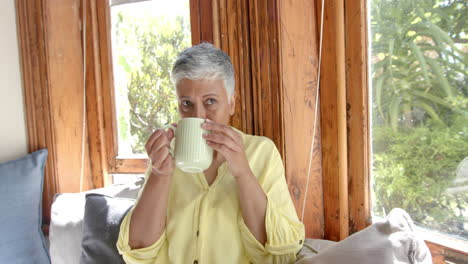Happy-senior-biracial-woman-sitting-on-couch-and-drinking-tea-at-home,-slow-motion