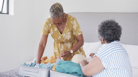 Happy-senior-african-american-female-friends-packing-suitcase-on-bed-at-holiday-home,-slow-motion