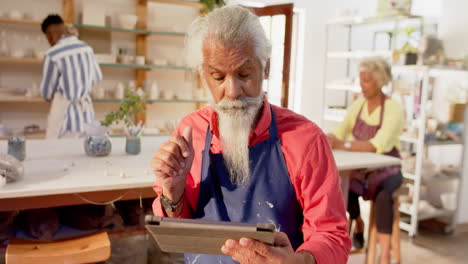 Focused-biracial-potter-with-long-beard-using-tablet-in-pottery-studio,-slow-motion