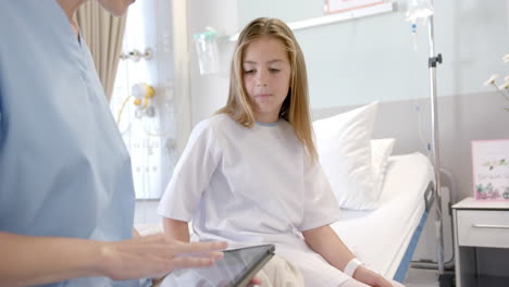 Caucasian-female-doctor-using-tablet-talking-with-girl-patient-sitting-on-hospital-bed,-slow-motion