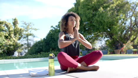 Happy-biracial-woman-practicing-yoga-meditation-sitting-by-pool-in-sunny-garden,-slow-motion