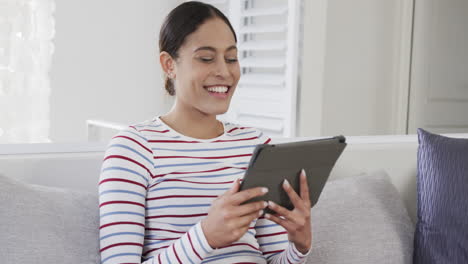 Happy-biracial-woman-sitting-on-sofa-using-tablet,-slow-motion