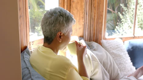 Glückliche-ältere-Frau-Mit-Gemischter-Abstammung,-Die-Auf-Der-Couch-Sitzt-Und-Zu-Hause-Tee-Trinkt,-Zeitlupe