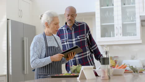 Happy-senior-biracial-couple-using-tablet-and-cooking-in-kitchen,-slow-motion