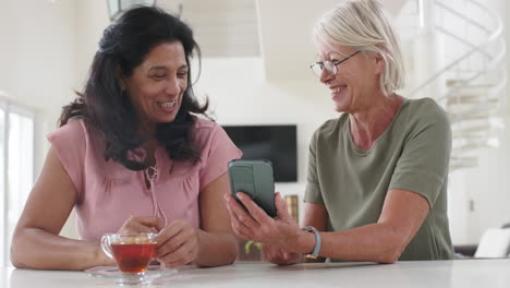 Happy-diverse-senior-women-using-smartphone-and-discussing-in-sunny-kitchen,-slow-motion