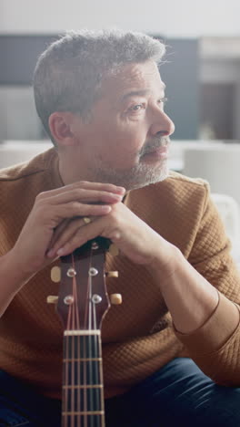 Vertical-video-of-happy-senior-biracial-man-sitting-on-couch-holding-guitar-at-home,-slow-motion