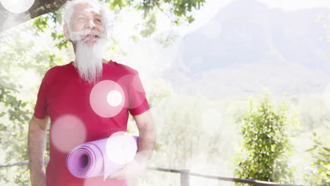 Animation-of-spots-of-light-and-trees-over-biracial-senior-man-with-yoga-mat-in-garden