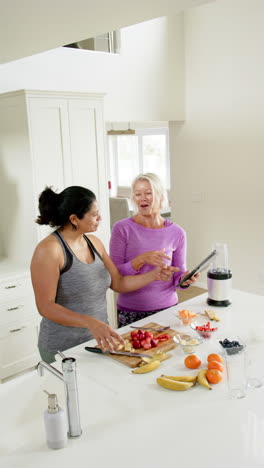Two-happy-diverse-senior-women-chopping-fruits-and-using-tablet-in-sunny-kitchen,-slow-motion