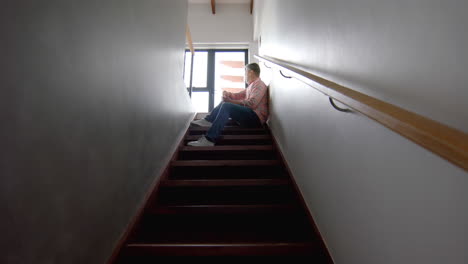 Thoughtful-senior-biracial-man-sitting-on-stairs-looking-out-of-window,-copy-space,-slow-motion
