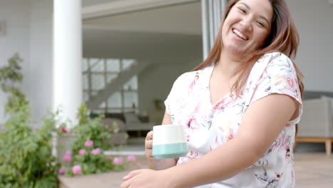 Plus-size-biracial-woman-sitting-on-terrace-drinking-coffee-and-laughing,-copy-space,-slow-motion