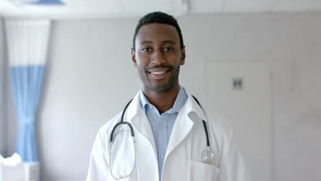 Portrait-of-happy-african-american-male-doctor-in-white-coat-smiling-in-hospital-ward,-slow-motion