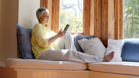 Glückliche-ältere-Frau-Mit-Gemischter-Abstammung,-Die-Auf-Der-Couch-Sitzt-Und-Zu-Hause-Ihr-Smartphone-Benutzt,-Zeitlupe