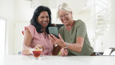Fröhliche,-Vielfältige-Seniorinnen,-Die-Smartphones-Benutzen-Und-In-Einer-Sonnigen-Küche-Diskutieren,-Zeitlupe