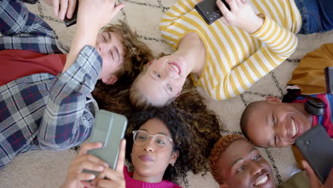 Happy-diverse-group-of-teenage-friends-using-smartphones-and-lying-on-carpet-at-home,-slow-motion