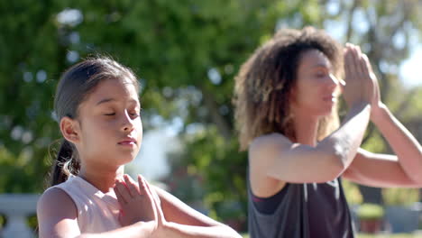Happy-biracial-mother-and-daughter-practicing-yoga-meditation-sitting-in-sunny-garden,-slow-motion