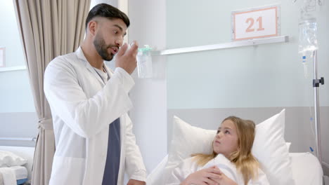 Diverse-male-doctor-showing-girl-patient-in-hospital-bed-how-to-use-asthma-inhaler,-slow-motion