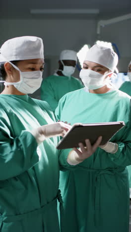 Vertical-video-of-diverse-female-surgeons-using-tablet-in-operating-theatre-at-hospital,-slow-motion