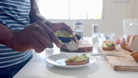 African-american-man-putting-avocado-on-toast-in-sunny-kitchen,-slow-motion