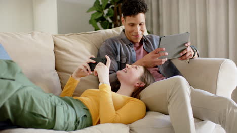 Happy-diverse-couple-sitting-on-sofa-using-smartphone-and-tablet-at-home,-in-slow-motion