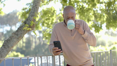 Happy-senior-biracial-man-at-balcony-using-smartphone-and-drinking-coffee-over-forest,-slow-motion