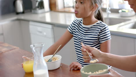 Biracial-mother-and-daughter-eating-breakfast-cereal-in-sunny-kitchen,-slow-motion
