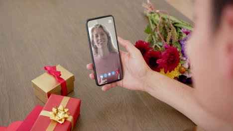 Biracial-woman-holding-smartphone-with-woman-talking-on-screen-with-gifts-on-desk
