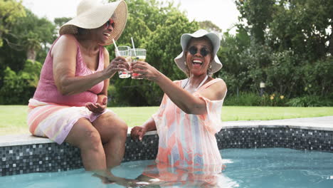 Happy-senior-african-american-female-friends-in-sunhats-sitting-in-pool-making-a-toast,-slow-motion