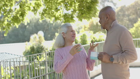 Happy-senior-biracial-couple-talking-and-drinking-coffee-at-balcony-at-home,-slow-motion