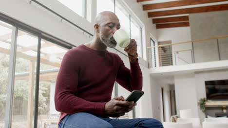 Happy-mature-biracial-man-drinking-coffee-and-using-smartphone-in-kitchen,-slow-motion