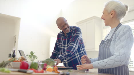 Feliz-Pareja-De-Ancianos-Diversos-Preparando-Verduras-Y-Usando-Tabletas-En-La-Cocina,-Cámara-Lenta
