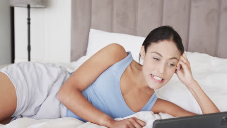 Happy-biracial-woman-lying-on-bed-using-tablet-for-video-call,-slow-motion