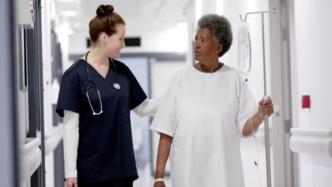 Happy-diverse-female-doctor-walking-with-senior-woman-with-drip-in-hospital-corridor,-slow-motion