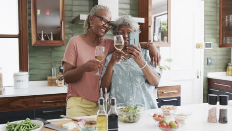 Happy-senior-african-american-female-friends-drinking-wine-taking-selfies-in-kitchen,-slow-motion