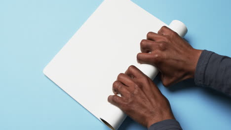 Video-Von-Händen-Eines-Afroamerikanischen-Mannes-Mit-Aufgerolltem-Weißem-Papier-Auf-Blauem-Hintergrund