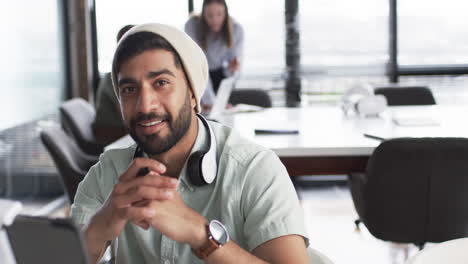 Young-Asian-man-with-a-beard-smiles-at-the-camera,-wearing-a-beanie-and-headphones-around-his-neck,-