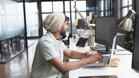 Young-Asian-man-works-on-laptop-in-a-modern-business-office-setting,-wearing-headphones-and-a-beanie
