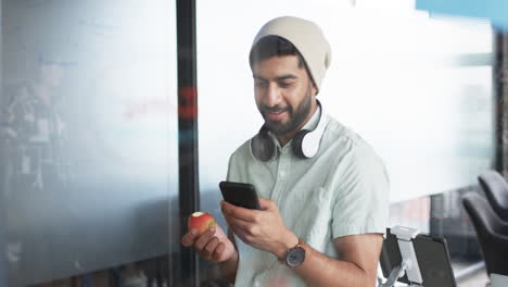Junger-Asiatischer-Geschäftsmann-Mit-Bart-Lächelt,-Während-Er-Sein-Smartphone-Benutzt-Und-Einen-Apfel-Hält