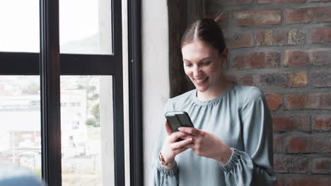 Junge-Kaukasische-Geschäftsfrau-Lächelt,-Während-Sie-Ihr-Smartphone-An-Einem-Fenster-Benutzt