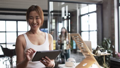 Asian-businesswoman-in-white-tank-top-holds-a-tablet,-smiling-at-the-camera