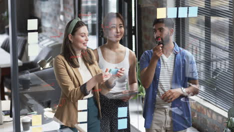 Young-Caucasian-woman-and-Asian-colleagues-brainstorm-with-sticky-notes-on-a-glass-wall-in-a-busines